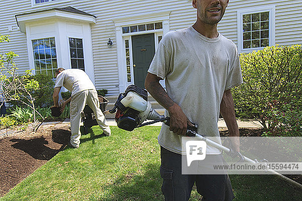 Landschaftsgärtner bei der Arbeit in einem Garten  einer beim Mulchen  der andere beim Kantenschneiden mit einem Kantenschneider