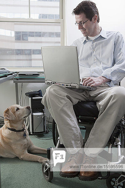 Ein Mann im Rollstuhl mit einer Rückenmarksverletzung arbeitet an einem Laptop  während ein Diensthund neben ihm sitzt