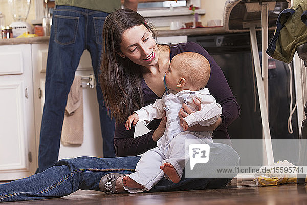 Mutter sitzt mit ihrem Sohn auf dem Boden  während der Vater in der Küche arbeitet