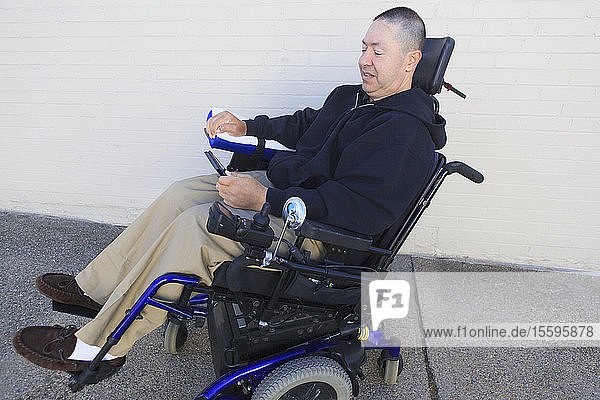 Mann mit Rückenmarksverletzung und nervengeschädigtem Arm im motorisierten Rollstuhl schaut auf sein Smartphone