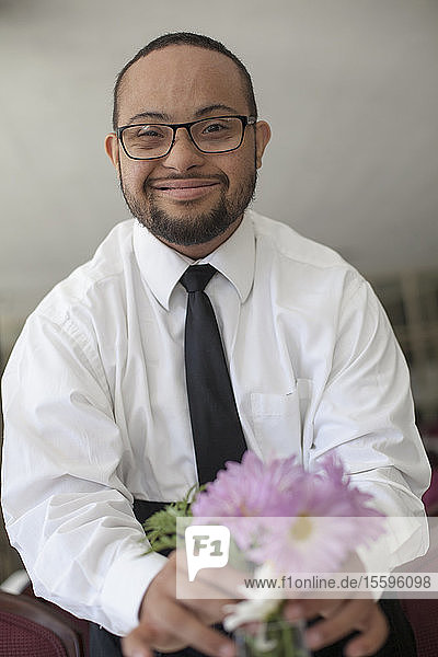 Porträt eines glücklichen afroamerikanischen Mannes mit Down-Syndrom  der als Kellner Blumen in einer Vase auf dem Tisch arrangiert