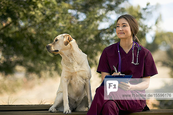 Lächelnde junge Krankenschwester sitzt zufrieden im sonnigen Garten mit einem freundlichen Hund