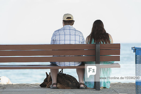 Ein blindes Paar sitzt mit seinem Diensthund auf einer Bank am Strand