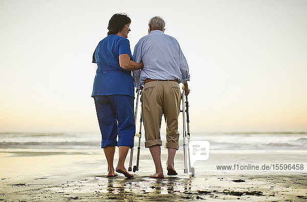 Ein älterer Mann geht mit einer Gehhilfe neben einer Krankenschwester am Strand