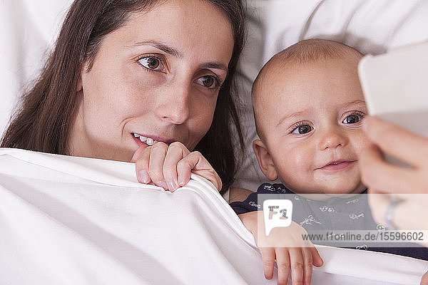 Nahaufnahme der Hand eines Mannes  der seiner Frau und seinem Baby im Bett ein Smartphone zeigt