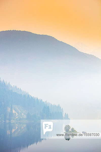 Dichter Nebel über einem See und einem Berg mit einem leuchtend orangefarbenen Himmel bei Sonnenaufgang; British Columbia  Kanada