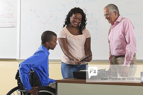 Two engineering students  one in wheelchair  looking at electronic tablet with Professor in a classroom