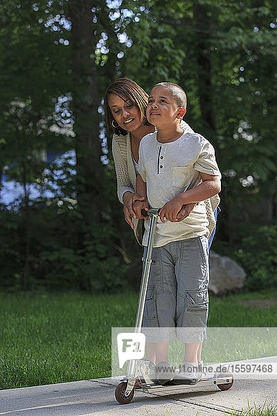 Hispanischer Junge mit Autismus  der mit seiner Mutter im Park auf einem Tretroller spielt