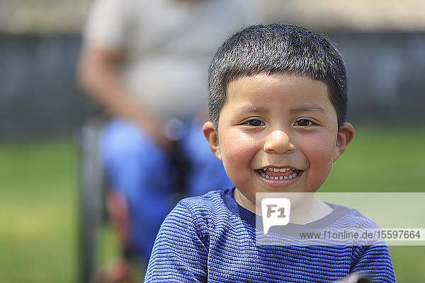 Glückliches Porträt eines hispanischen Kindes mit seinem Vater im Rollstuhl mit Rückenmarksverletzung im Hintergrund