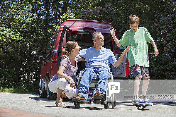 Mann mit Rückenmarksverletzung im Rollstuhl gibt seinem Sohn auf dem Skateboard ein High-Five