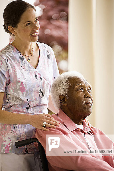 Eine lächelnde Krankenschwester legt einem älteren Mann  der in einem Rollstuhl auf einer Veranda sitzt  unterstützend die Hand auf die Schultern