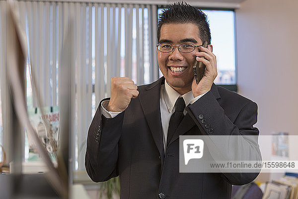 Fröhlicher asiatischer Mann mit Autismus  der mit seinem Handy in einem Büro jubelt