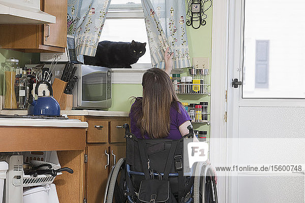 Frau mit Spina Bifida im Rollstuhl spielt mit einer Katze im Küchenfenster