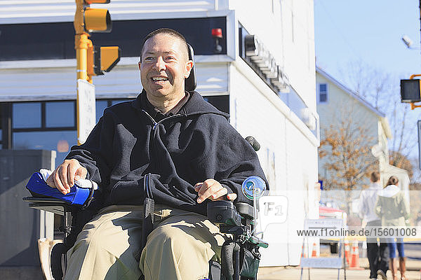 Mann mit Rückenmarksverletzung und Arm mit Nervenschaden im motorisierten Rollstuhl überquert beim Einkaufen eine öffentliche Straße