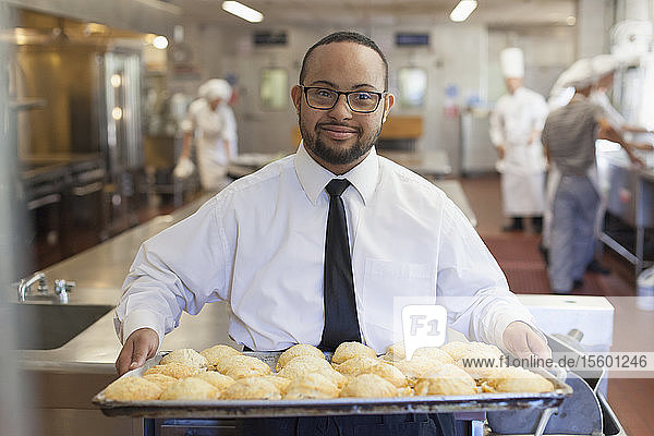 Afroamerikanischer Mann mit Down-Syndrom als Koch mit einem Tablett voller Kekse in einer Großküche