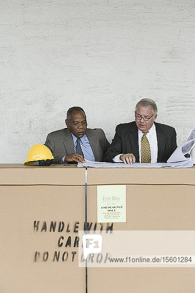Zwei Geschäftsleute sitzen an einer Pappschachtel und diskutieren über einen Bauplan