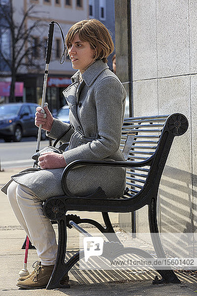 Junge blinde Frau mit ihrem Blindenstock auf einer Stadtbank sitzend