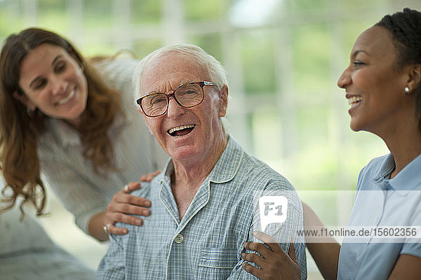 Porträt eines lachenden älteren Mannes  der von einer Krankenschwester und seiner erwachsenen Tochter getröstet wird  während er einen Pyjama trägt.