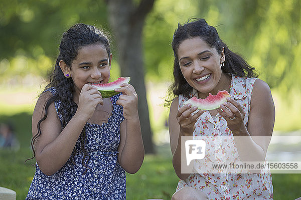 Happy Hispanic mother with her teen daughter eating watermelon in park