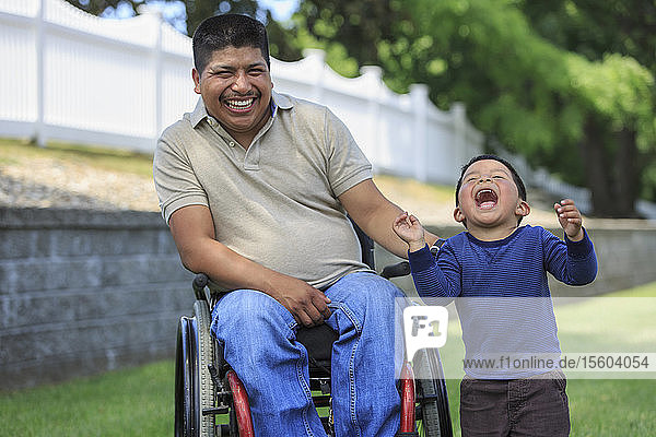 Hispanischer Mann mit Rückenmarksverletzung im Rollstuhl mit seinem Sohn lachend auf dem Rasen
