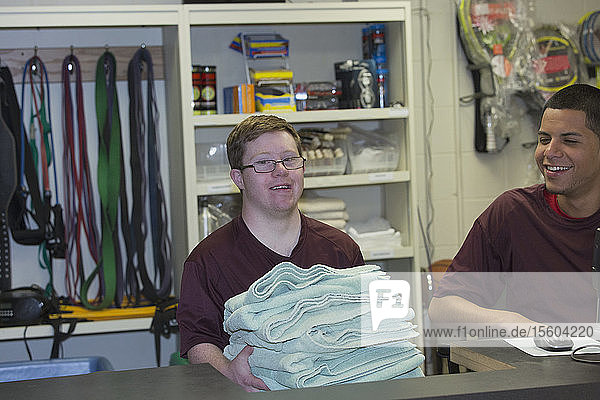 Junger Mann mit Down-Syndrom bereitet Handtücher in der Ausrüstungsabteilung des Colleges für das Fitnessstudio mit einem Betreuer vor