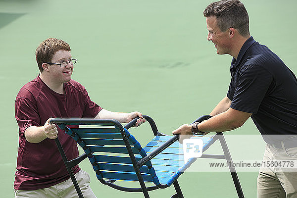 Junger Mann mit Down-Syndrom  der zusammen mit einem Betreuer Möbel auf einem College-Ballplatz transportiert