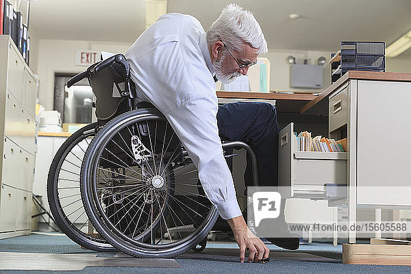 Man with Muscular Dystrophy in a wheelchair reaching for something he dropped at his office desk