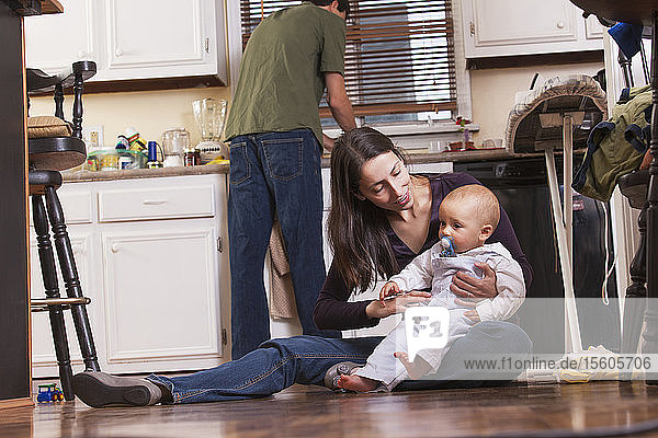 Mutter sitzt mit ihrem Sohn auf dem Boden  während der Vater in der Küche arbeitet