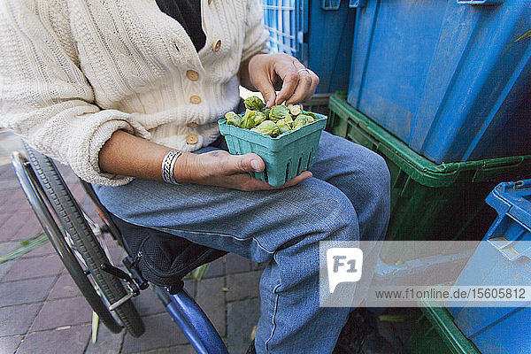 Frau mit Querschnittslähmung sitzt im Rollstuhl und kauft auf einem Markt Rosenkohl ein