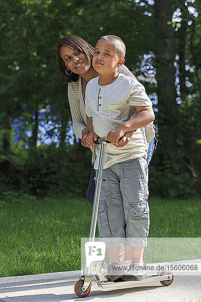 Hispanischer Junge mit Autismus  der mit seiner Mutter im Park auf einem Tretroller spielt