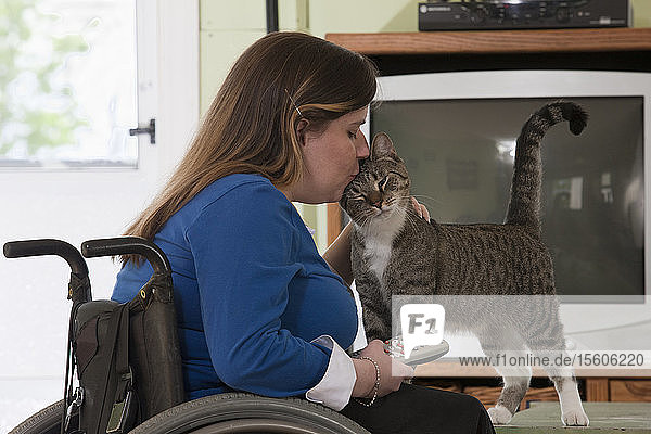 Frau mit Spina Bifida im Rollstuhl  die eine Katze küsst  während sie die Fernbedienung des Fernsehers hält