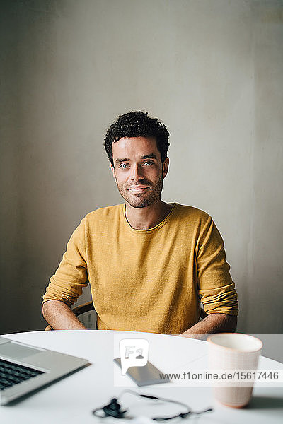 Portrait of confident mid adult man sitting at table against wall
