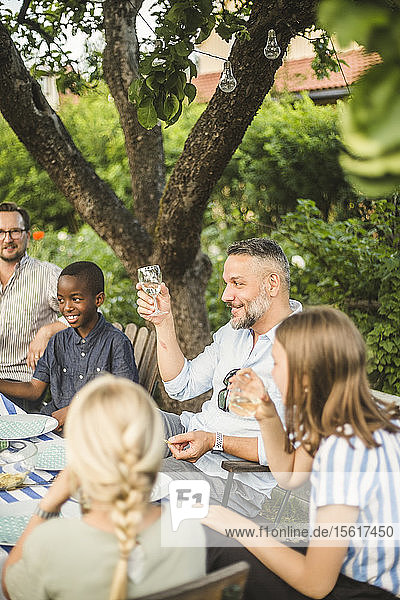 Reifer Mann hebt ein Weinglas  während er mit einem Freund und Kindern im Hinterhof sitzt