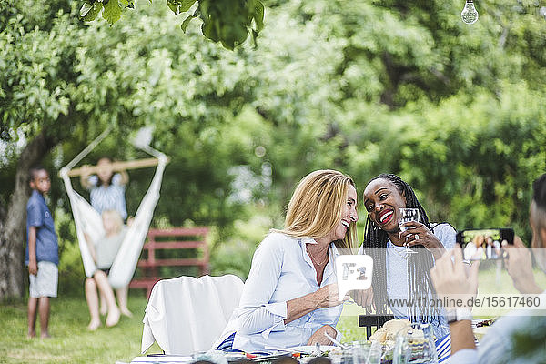 Mann mit Handy fotografiert lächelnde Frauen  die am Wochenende Wein im Garten feiern