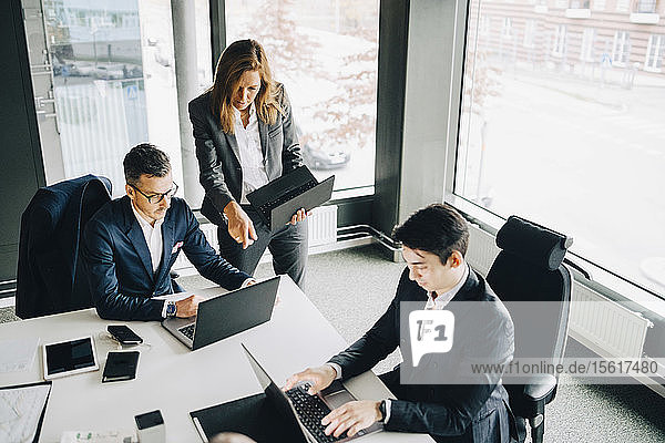Berufstätige Frau diskutiert mit einem Kollegen über einen Laptop  während ein Geschäftsmann am Konferenztisch im Büro arbeitet