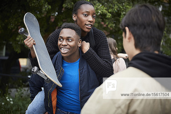 Lächelnder Teenager Junge  der eine Freundin  die ein Skateboard hält  huckepack nimmt
