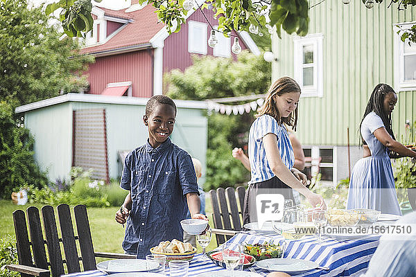 Lächelnder Junge mit Schüssel steht neben Mädchen am Esstisch bei einer Party im Hinterhof