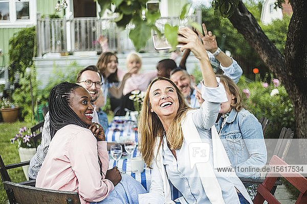 Happy woman taking selfie with friends on smart phone in backyard during garden party