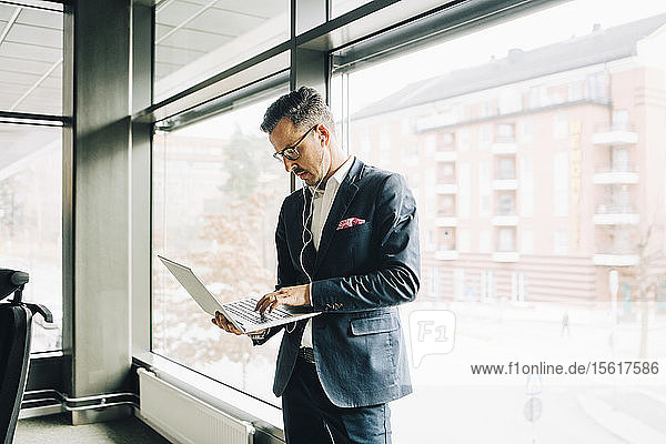 Mature businessman using laptop while standing against window in office