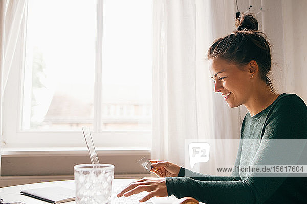 Side view of smiling woman with credit card using laptop in living room