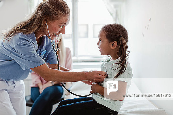 Weibliche Kinderärztin untersucht den Herzschlag des Mädchens durch ein Stethoskop  während die Mutter im Hintergrund in der Klinik sitzt
