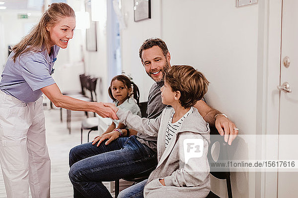 Smiling female pediatrician handshaking with boy sitting with family in hospital corridor