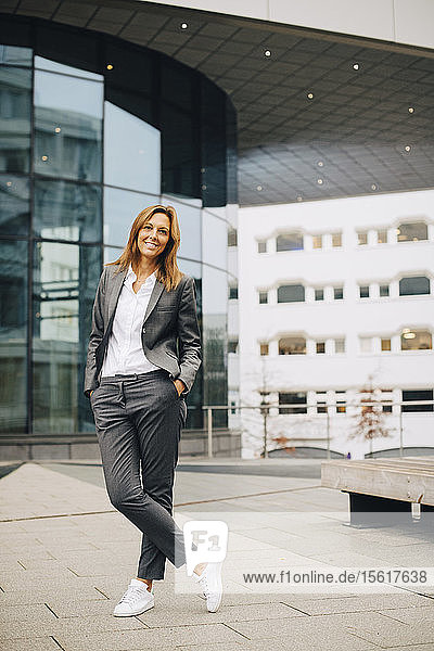 Selbstbewusst lächelnde Geschäftsfrau in voller Länge an Glasgebäude stehend