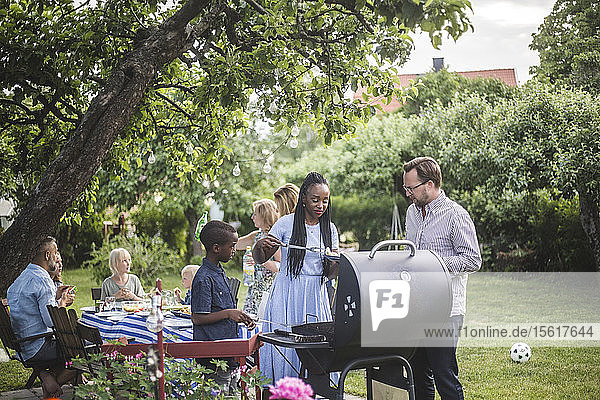Man talking to woman with son preparing food at barbecue grill against friends during weekend party