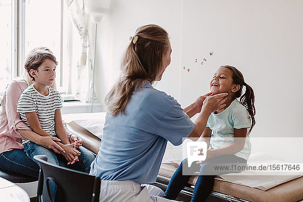 Weibliche Ärztin untersucht lächelndes Mädchen  während die Familie im Behandlungszimmer sitzt