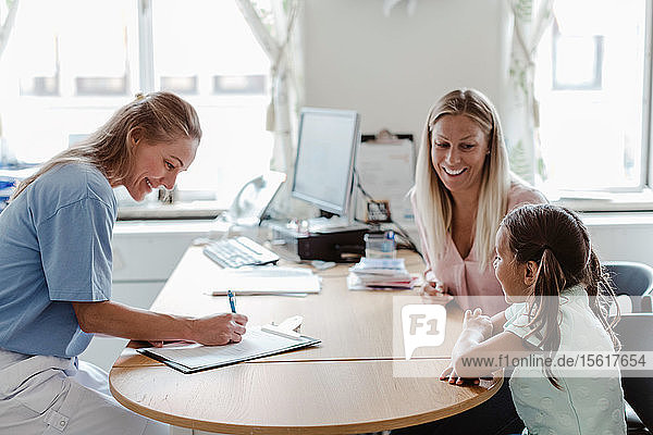 Lächelnder Kinderarzt schreibt ein Rezept  während ein Mädchen mit der Mutter am Schreibtisch in der Klinik sitzt