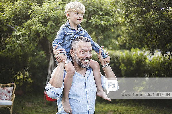 Smiling mature man carrying son on shoulder while having fun in backyard
