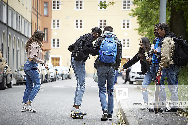 Teenage boy supporting female skateboarding while friends playing with ball on street in city