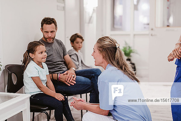 Weibliche Kinderärztin im Gespräch mit einem Mädchen  das im Krankenhauskorridor bei der Familie sitzt