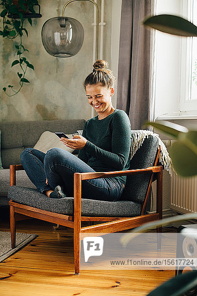Mittlere erwachsene Frau  die lächelt  während sie zu Hause ein Mobiltelefon auf einem Sessel benutzt